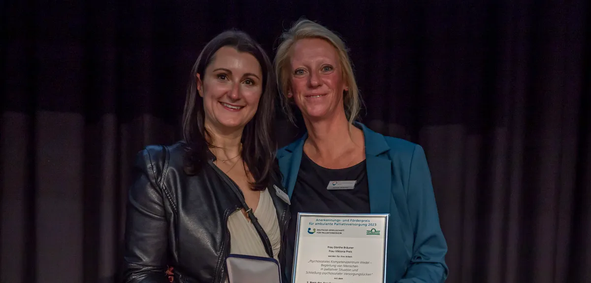 Dörthe Bräuner und Viktoria Preis stehen auf einer Bühne und halten die Urkunde des Anerkennungs- und Förderpreises für ambulante Palliativversorgung 2023.