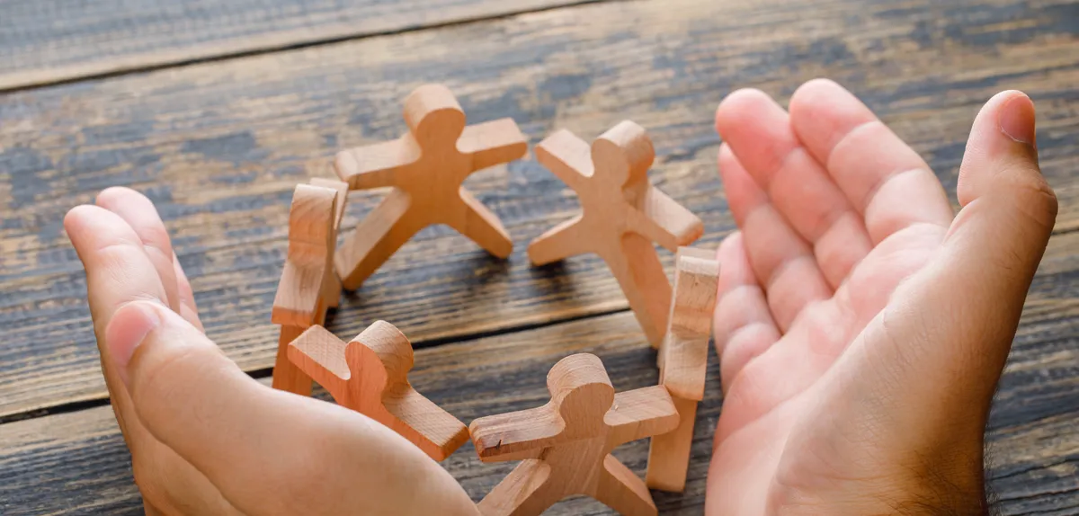 Zwei geöffnete Hände umschließen einen Kreis aus Holzfiguren, die sich an den Händen halten.