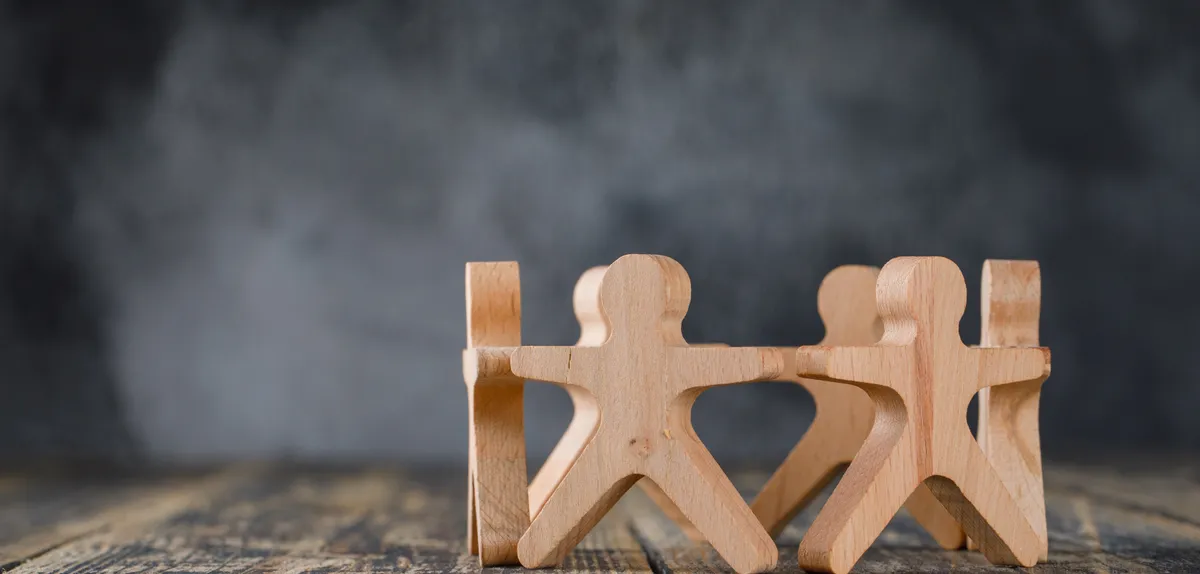 Sechs Holzfiguren stehen nebeneinander auf einer Holzfläche und halten sich an den Händen, vor dunklem Grund.